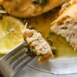 A fork holding up one bite of lemon caper chicken over a dish of more.