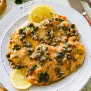 Two breasts of lemon caper chicken on a dish garnished with lemon slices.