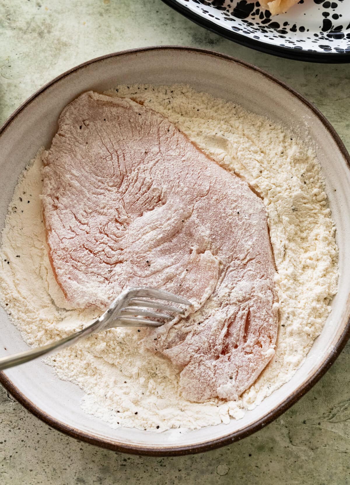 Chicken breast in a bowl of flour with a fork covering it. 