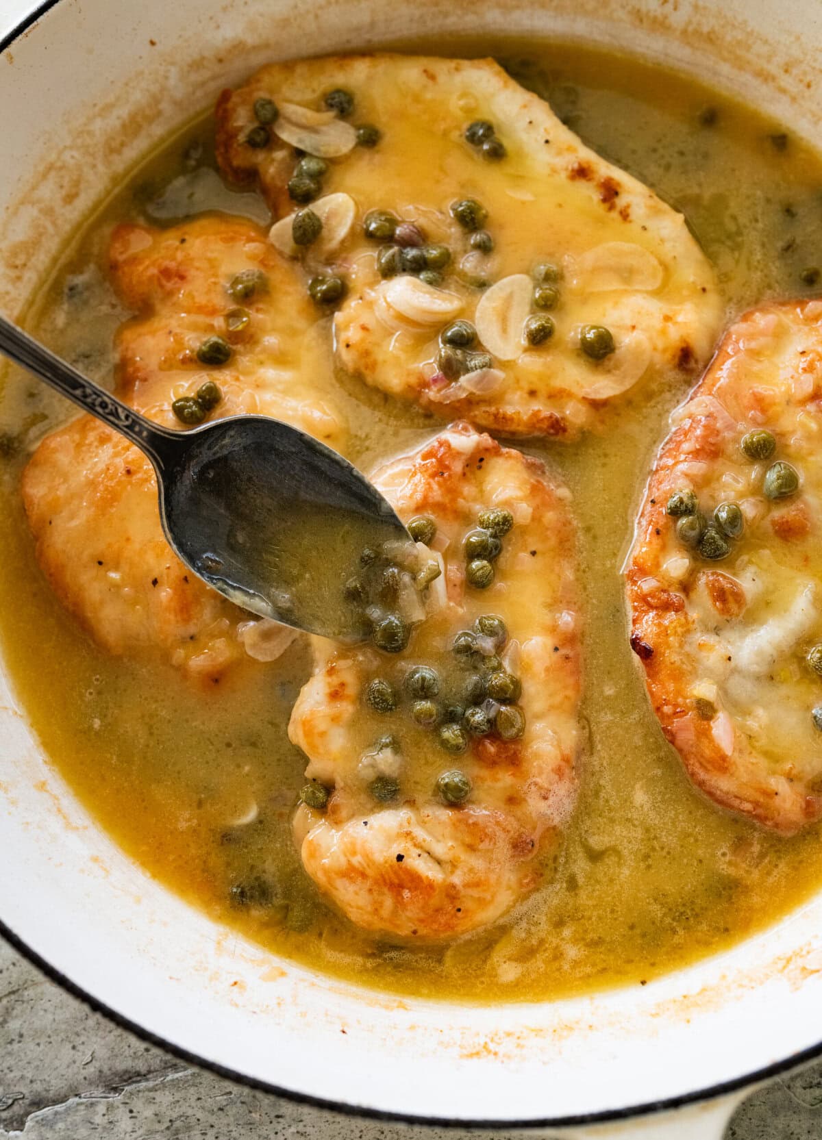 Capers and chicken added to the skillet, with a spoon pouring sauce over each breast. 