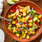 a bowl of mango salsa with a spoon resting in it and other ingredients to make the recipe nearby.
