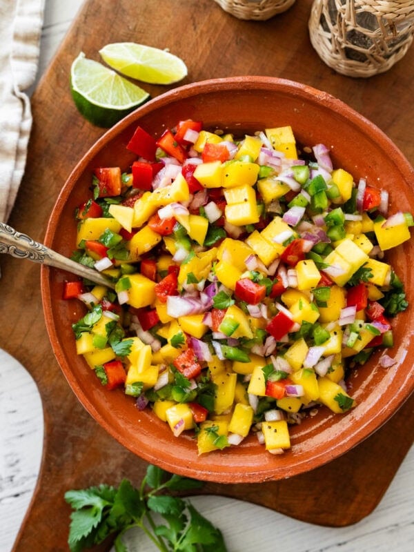 a bowl of mango salsa with a spoon resting in it and other ingredients to make the recipe nearby.