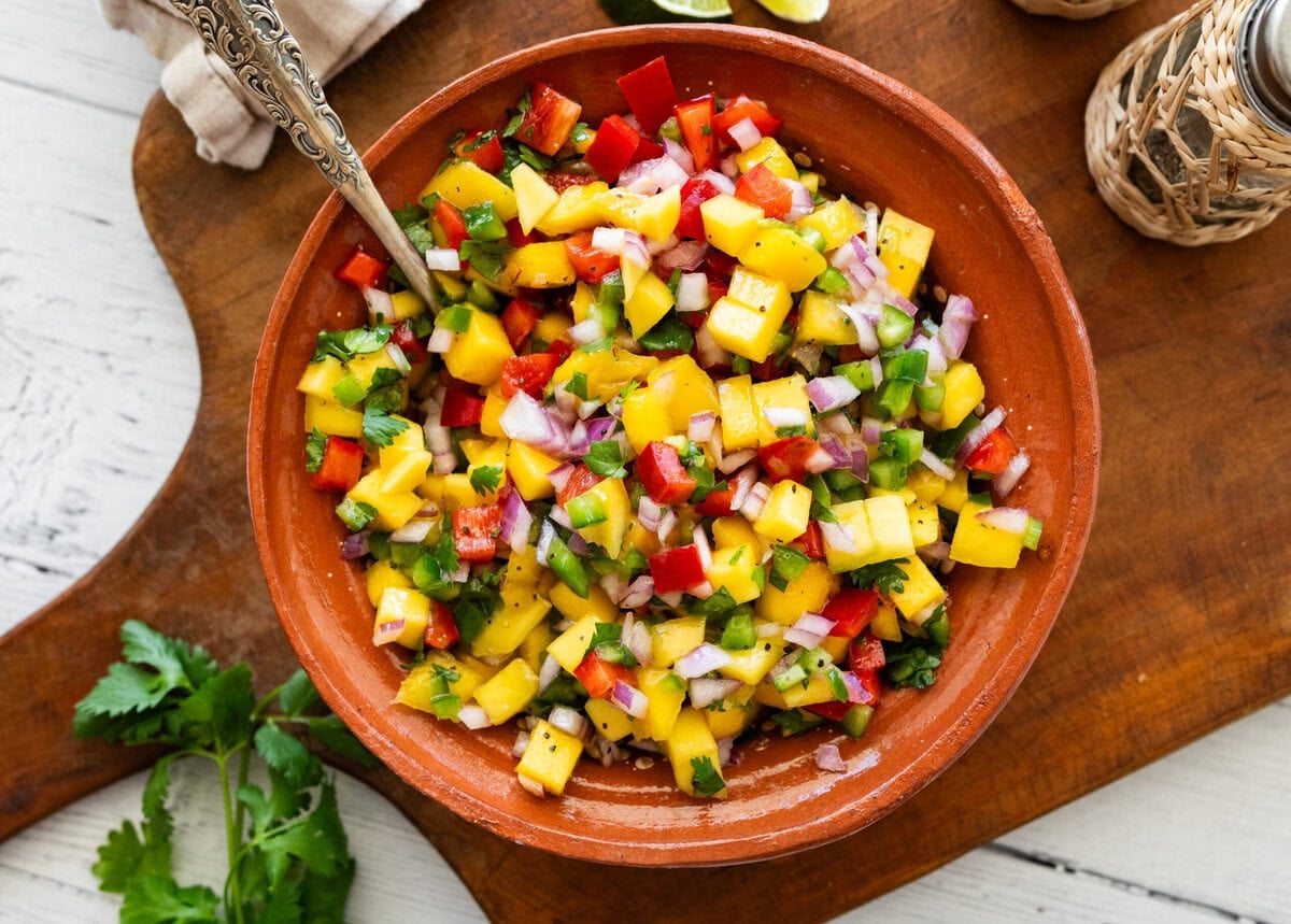 a bowl of mango salsa with a spoon resting in it and other ingredients to make the recipe nearby.