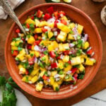 a bowl of mango salsa with a spoon resting in it and other ingredients to make the recipe nearby.