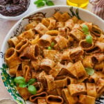A bowl of sun-dried tomato pesto pasta with garnish, and more ingredients nearby.