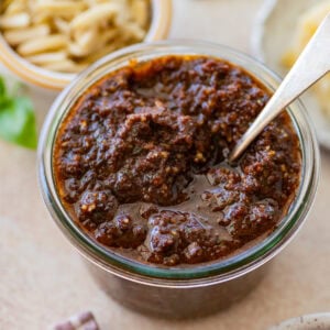 Sun-dried tomato pesto in a bowl with a spoon resting inside, and more ingredients nearby.