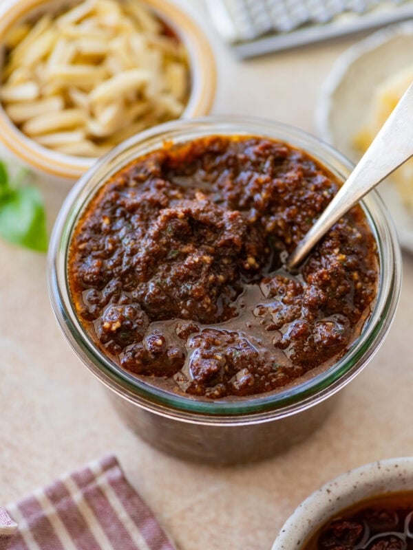 Sun-dried tomato pesto in a bowl with a spoon resting inside, and more ingredients nearby.