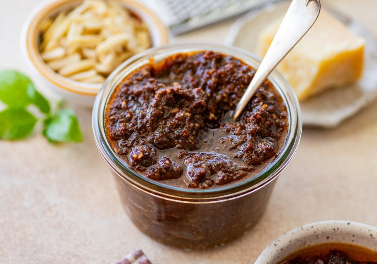 Sun-dried tomato pesto in a bowl with a spoon resting inside, and more ingredients nearby.