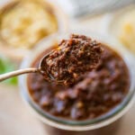Sun-dried tomato pesto in a bowl with a spoon holding up some, and more ingredients nearby.