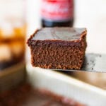A slice of coca cola cake resting on a serving utensil with more ingredients nearby.