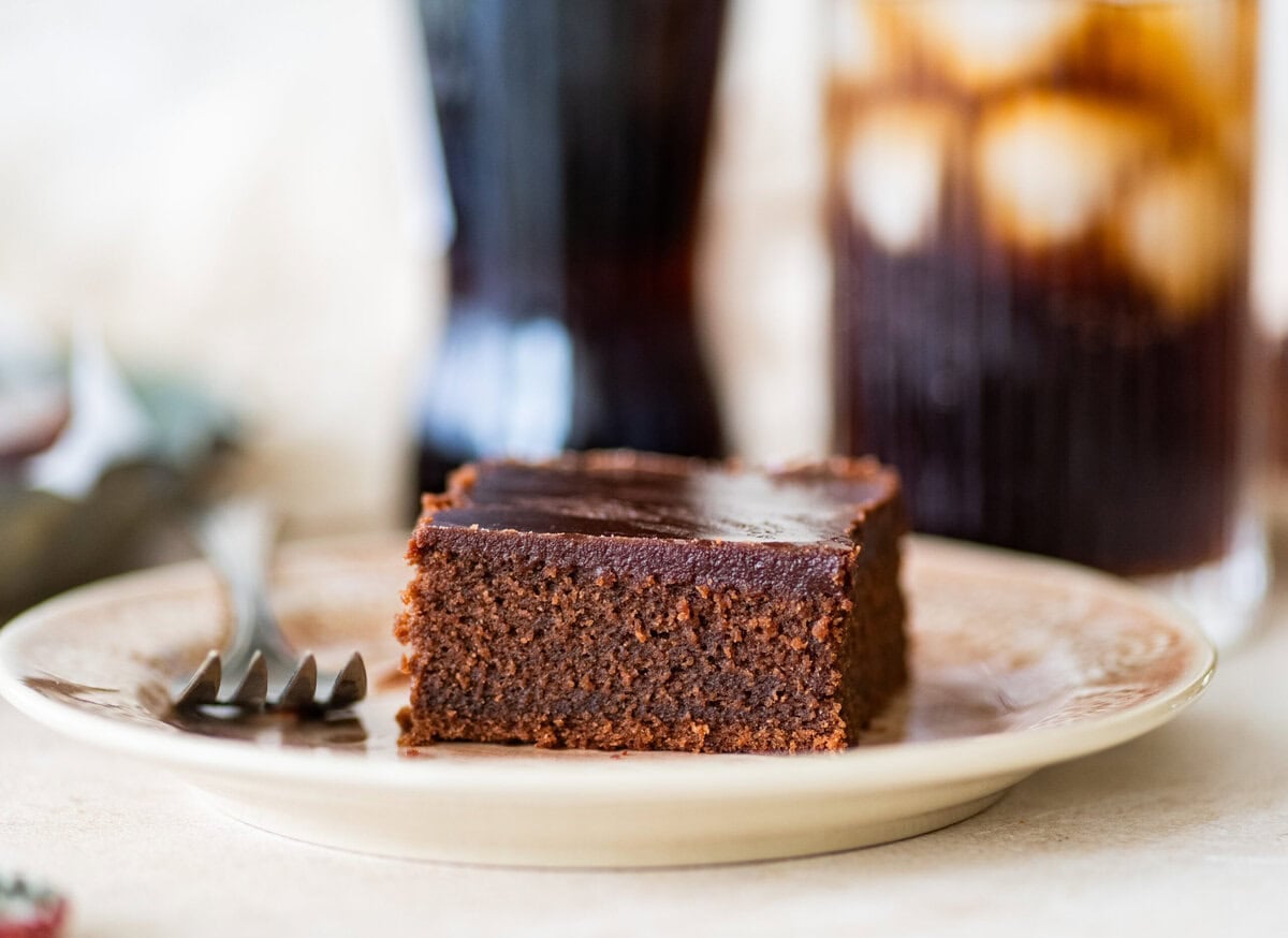 A slice of coca cola cake on a plate with a fork, with more ingredients nearby.