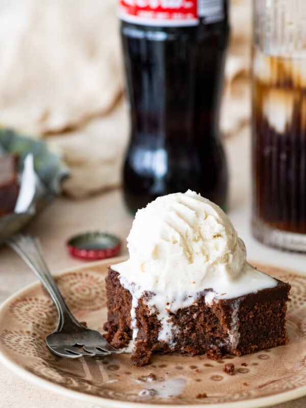 A slice of coca cola cake on a plate with a fork, topped with a scoop of ice cream with more ingredients nearby.