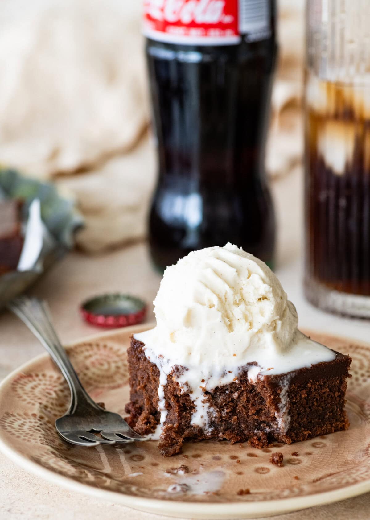 A slice of coca cola cake on a plate with a fork, topped with a scoop of ice cream with more ingredients nearby.