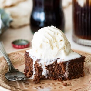 A slice of coca cola cake on a plate with a fork, topped with a scoop of ice cream with more ingredients nearby.