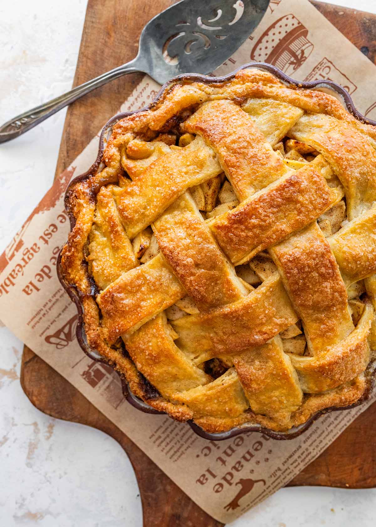 baked lattice-top apple pie on a newspaper-lined wooden cutting board with a pie server.