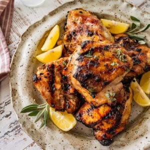 grilled chicken thighs with lemon wedges and fresh sage sprigs on a white platter.