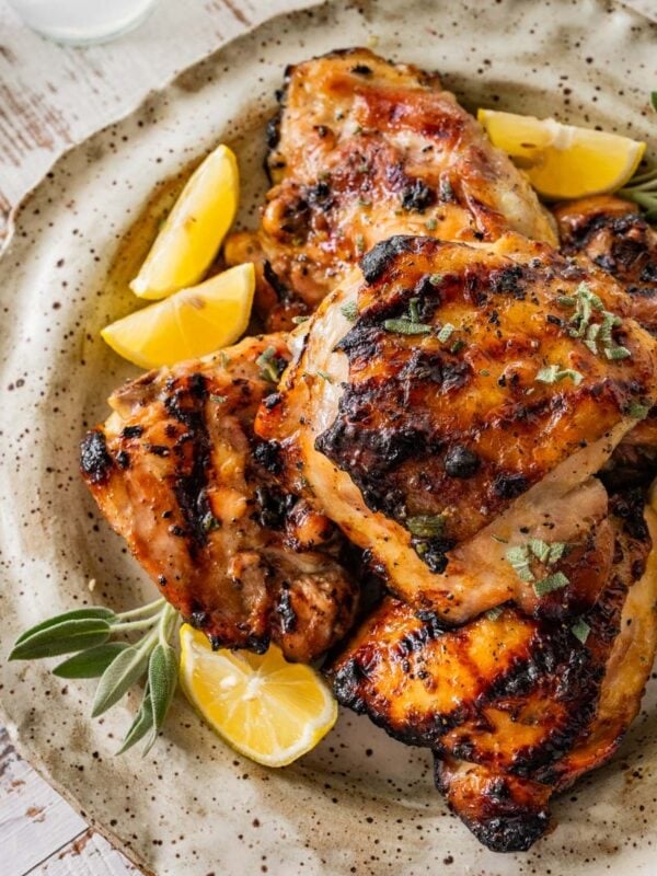 grilled chicken thighs with lemon wedges and fresh sage sprigs on a white platter.