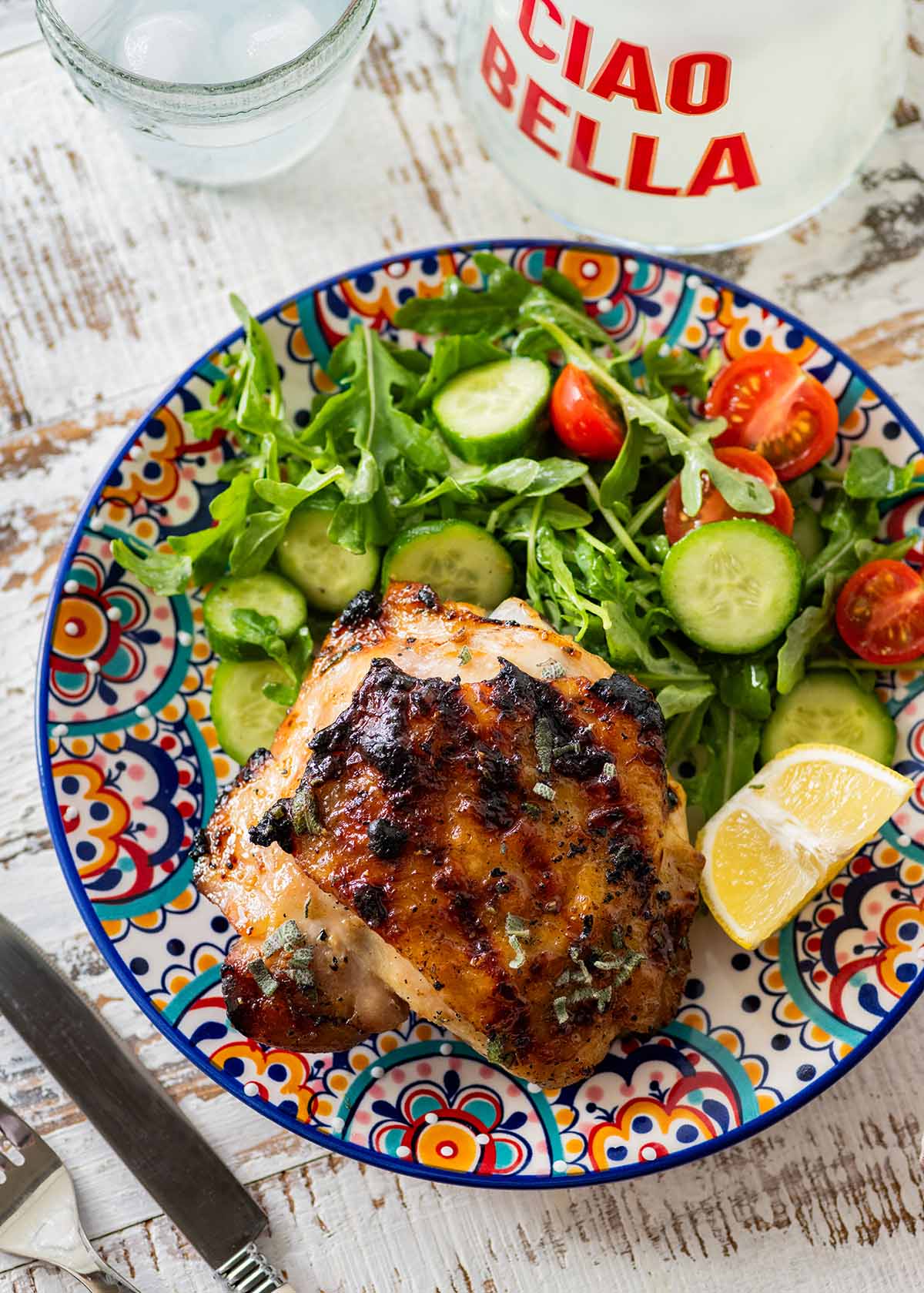 grilled chicken thigh on a colorful plate with a glass of water and side salad.