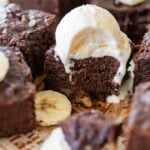 Sliced banana brownies on parchment paper with some banana slices around them. One brownie is topped with a scoop of ice cream and has a spoon removing a bite.