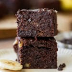 Two banana brownies stacked on top of each other on a plate with sliced bananas nearby.