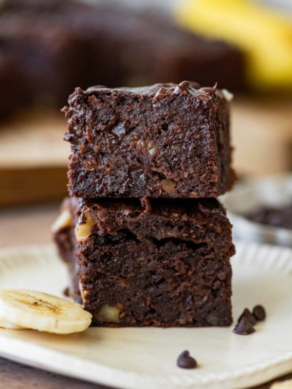Two banana brownies stacked on top of each other on a plate with sliced bananas nearby.