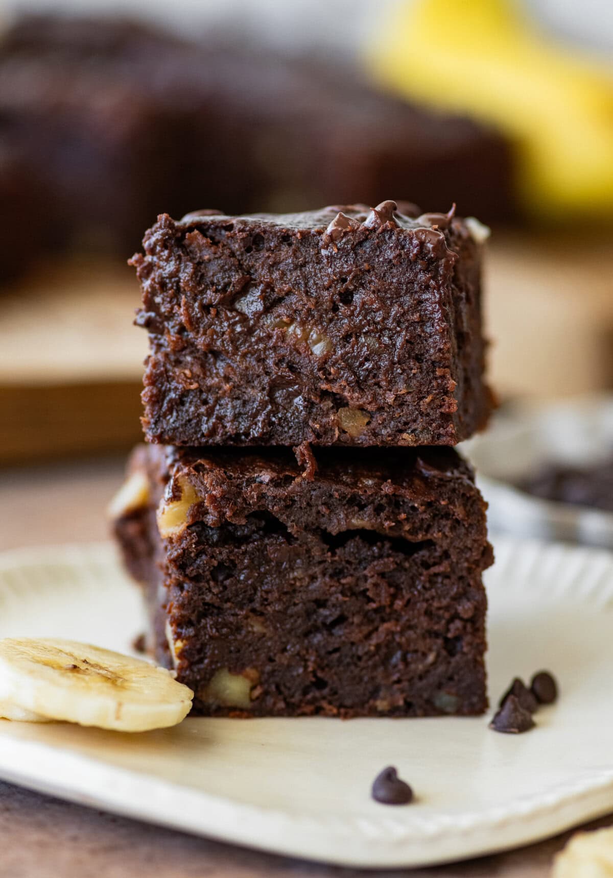 Two banana brownies stacked on top of each other on a plate with sliced bananas nearby.