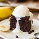 A banana brownie on a plate with a scoop of vanilla ice cream on top, sliced bananas nearby, and more ingredients in the background.