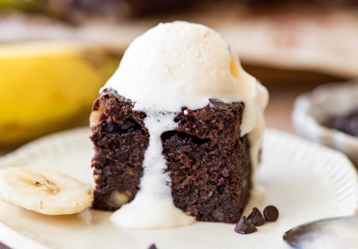 A banana brownie on a plate with a scoop of vanilla ice cream on top, sliced bananas nearby, and more ingredients in the background.