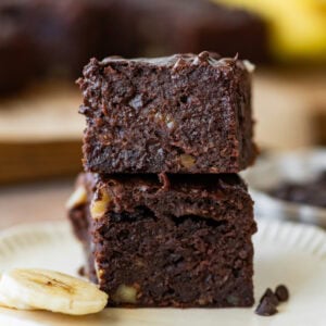 Two banana brownies stacked on top of each other on a plate with sliced bananas nearby.