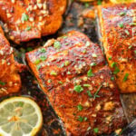 Blackened salmon in a skillet with parsley as a garnish and lemon slices scattered around each piece.