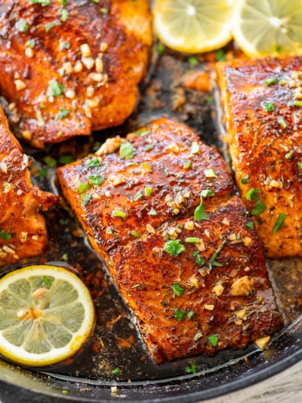 Blackened salmon in a skillet with parsley as a garnish and lemon slices scattered around each piece.