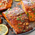 Blackened salmon in a skillet with parsley as a garnish and lemon slices scattered around each piece.