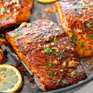 Blackened salmon in a skillet with parsley as a garnish and lemon slices scattered around each piece.