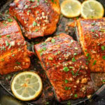 Blackened salmon in a skillet with parsley as a garnish and lemon slices scattered around each piece.
