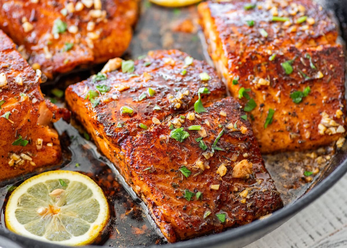 Blackened salmon in a skillet with parsley as a garnish and lemon slices scattered around each piece.