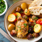 A plate of chicken scarpariello with a piece of bread resting on top and more ingredients nearby.