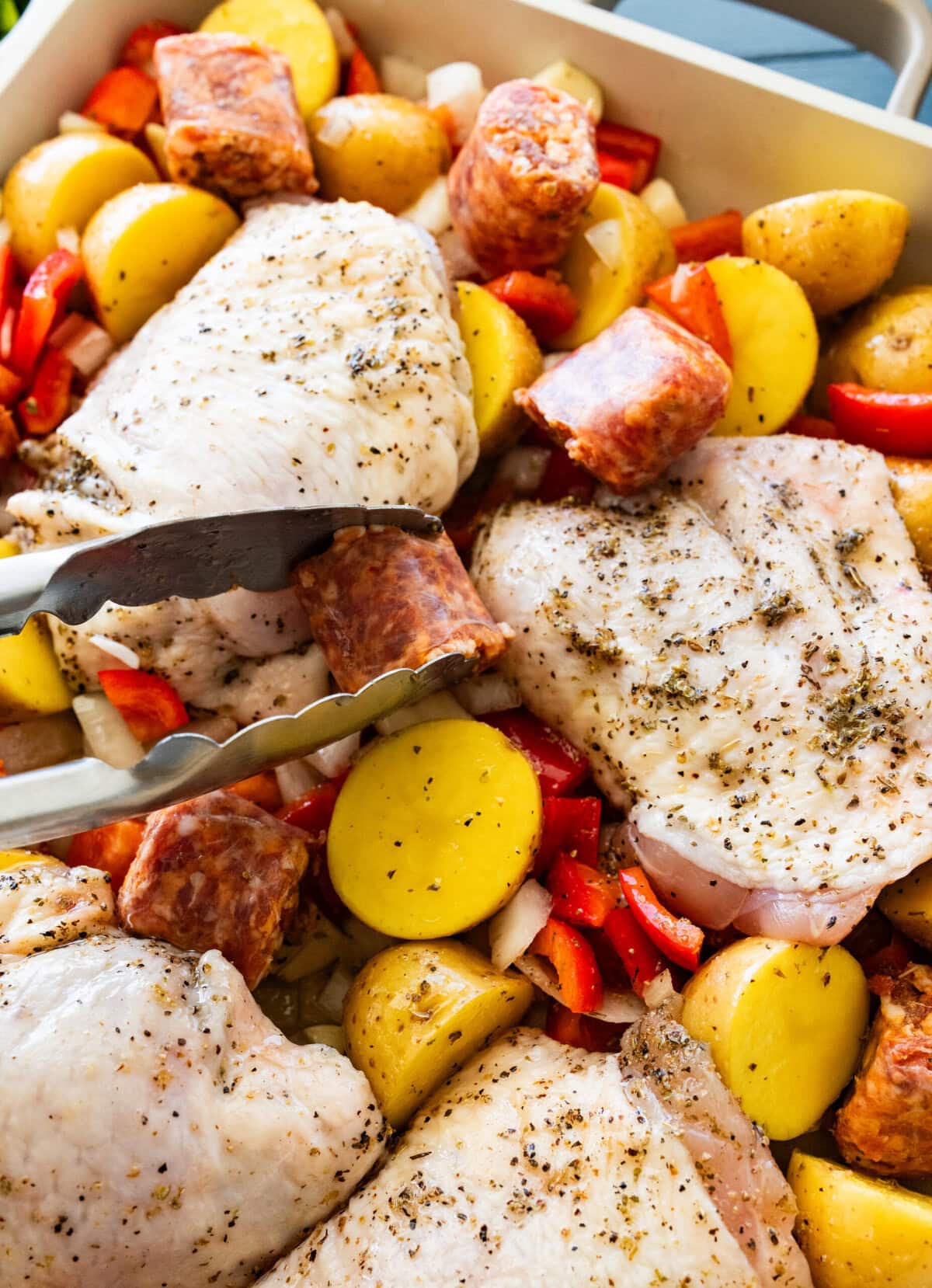 Tongs placing the sausage pieces around the chicken in a baking dish.