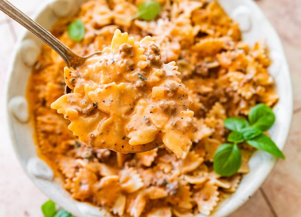 A large bowl of beef and bowtie pasta with a smaller bowl nearby, topped with basil as garnish.