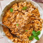 A large bowl of beef and bowtie pasta with a smaller bowl nearby, topped with basil and a spoon resting inside,