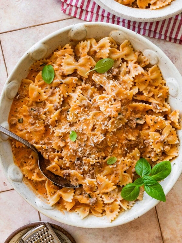 A large bowl of beef and bowtie pasta with a smaller bowl nearby, topped with basil and a spoon resting inside,