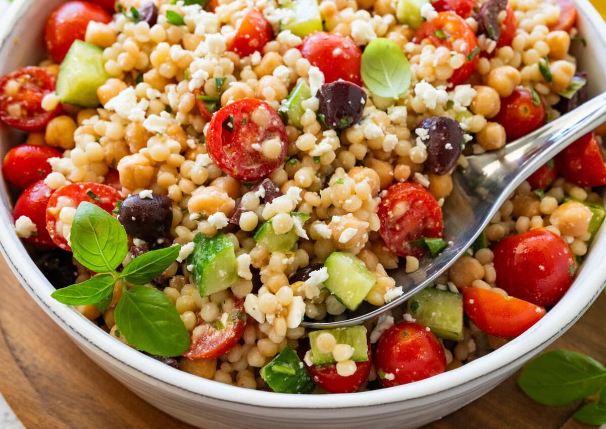 Pearl couscous salad in a serving bowl with a spoon lifting up some, and more ingredients nearby.