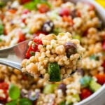 Pearl couscous salad in a serving bowl with a spoon lifting up some, and more ingredients nearby.