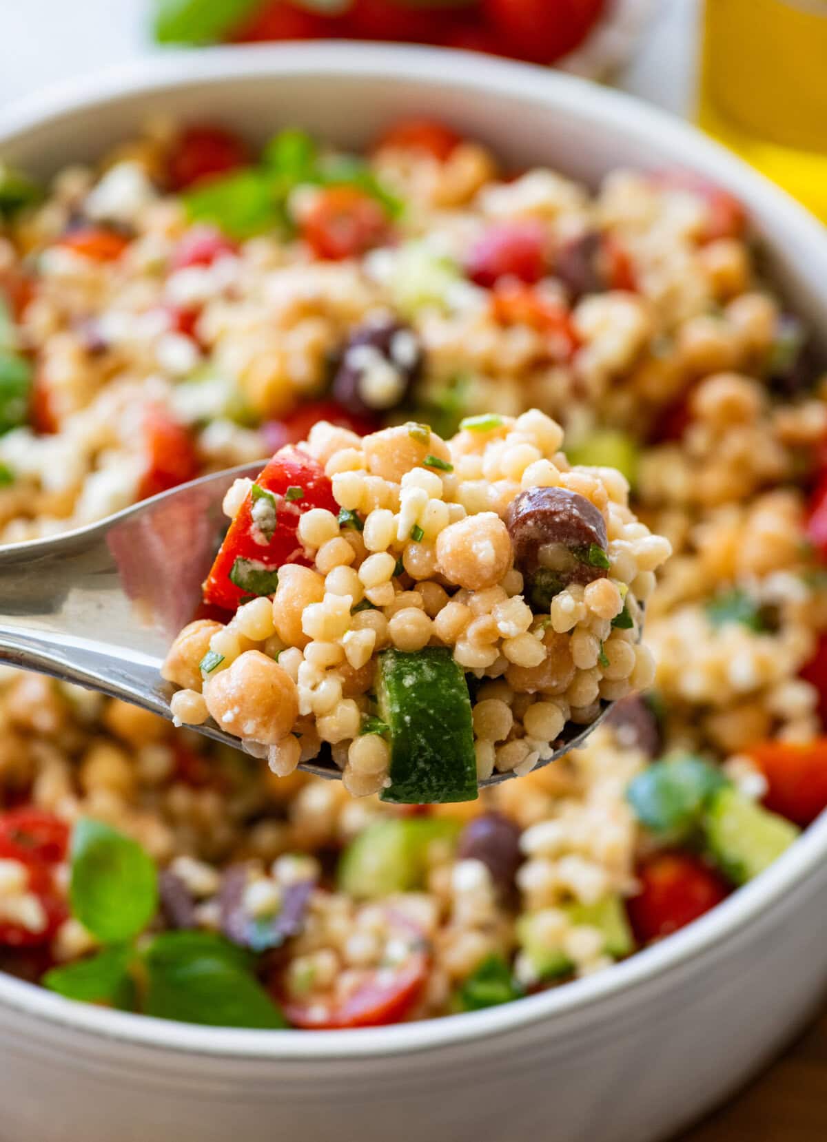 Pearl couscous salad in a serving bowl with a spoon lifting up some, and more ingredients nearby.