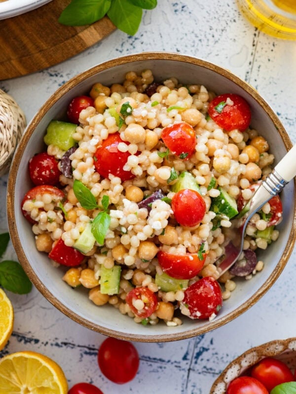 Pearl couscous salad in a serving bowl with a spoon and more ingredients nearby.