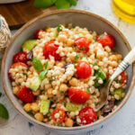 Pearl couscous salad in a serving bowl with a spoon lifting up some, and more ingredients nearby.