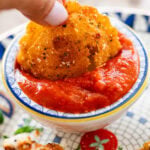 Dipping one fried ravioli into a bowl of marinara sauce with more ravioli nearby.