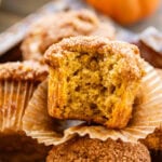 A tray of pumpkin banana muffins, with one half unwrapped with a bite taken out, with bananas and pumpkins nearby.