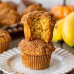 Two muffins stacked on top of each other on a plate, with the top muffin having a bite taken out, and more muffins, bananas, and pumpkins nearby.