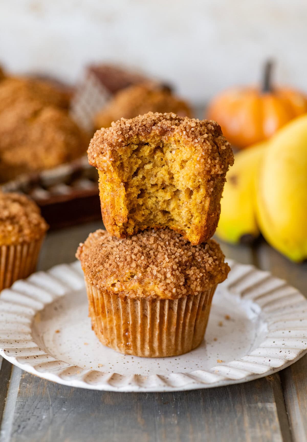 Two muffins stacked on top of each other on a plate, with the top muffin having a bite taken out, and more muffins, bananas, and pumpkins nearby.