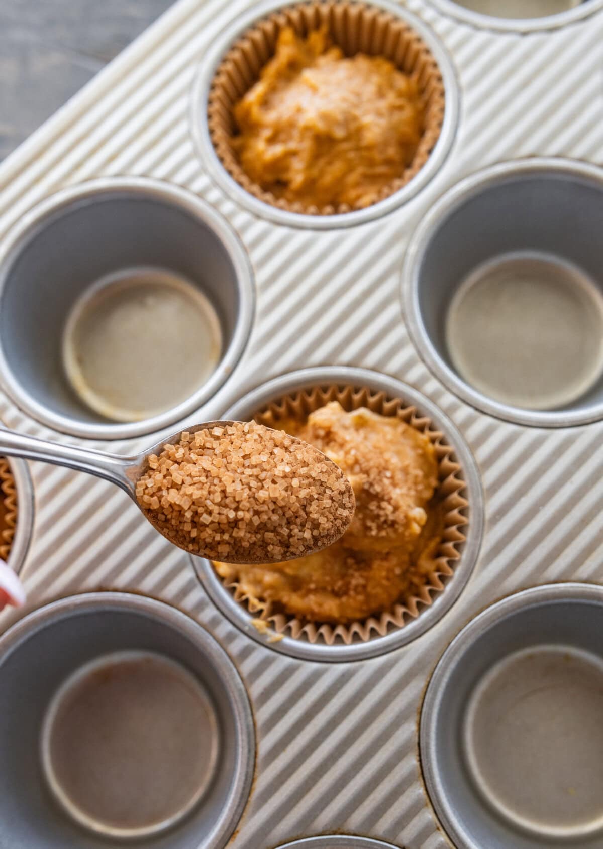 Sugar and cinnamon mix being spooned on top of muffin batter in a muffin tin.